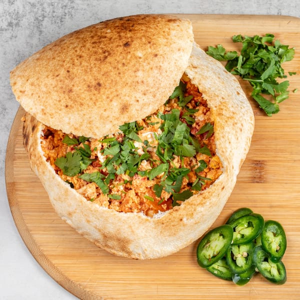 Baked pizza dough bowl filled with scrambled eggs, cheese, and cilantro, with jalapeños on the side.