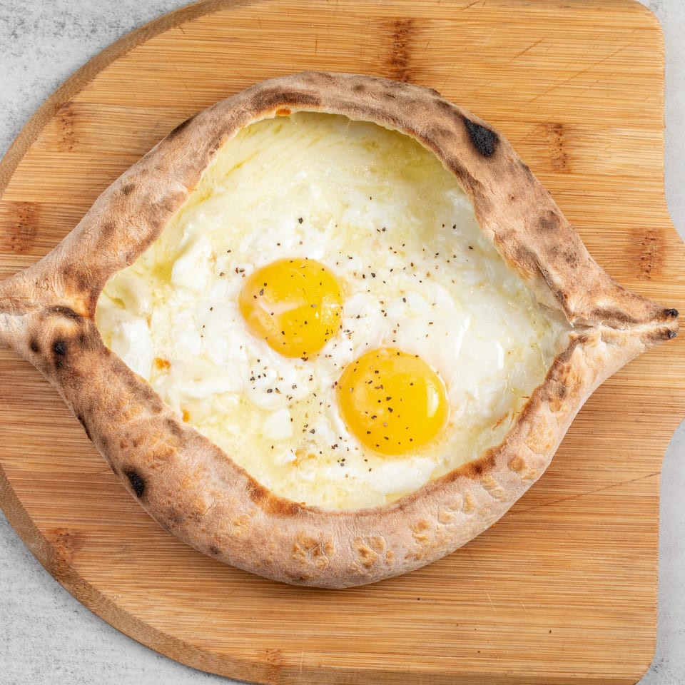 Baked Pizza and Sandwiches' boat-shaped pizza with melted cheese, two sunny-side-up eggs, and pepper.
