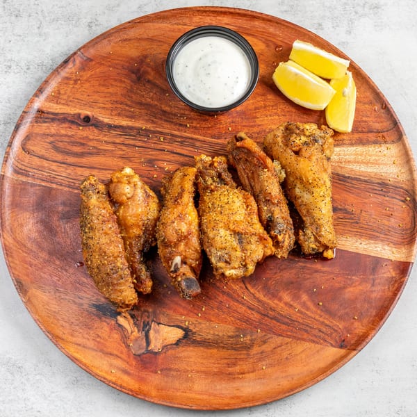 Crispy chicken wings seasoned with pepper, served on a wooden platter with ranch and lemon wedges.