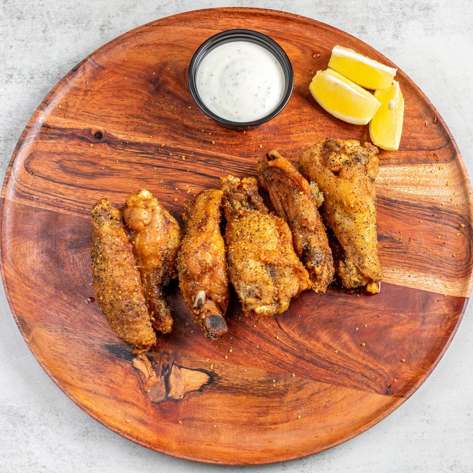 Crispy chicken wings seasoned with pepper, served on a wooden platter with ranch and lemon wedges.