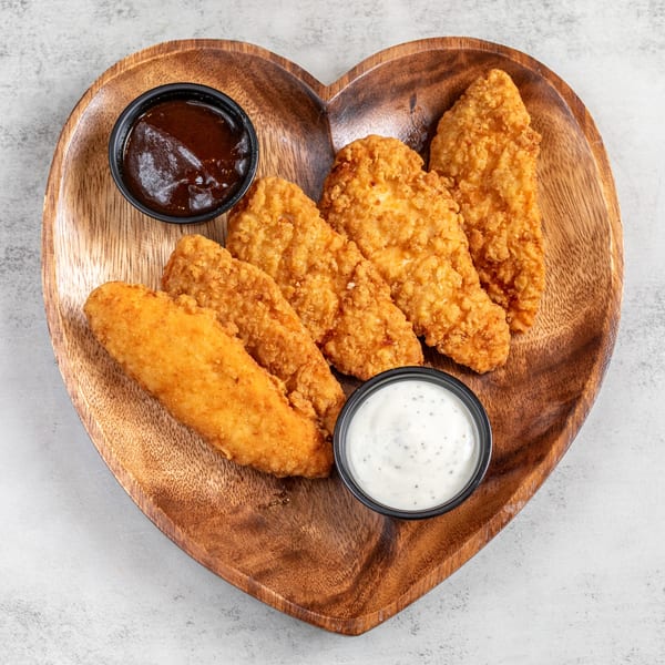 Golden-brown fried chicken tenders with barbecue and ranch dipping sauces on a heart-shaped wooden board.